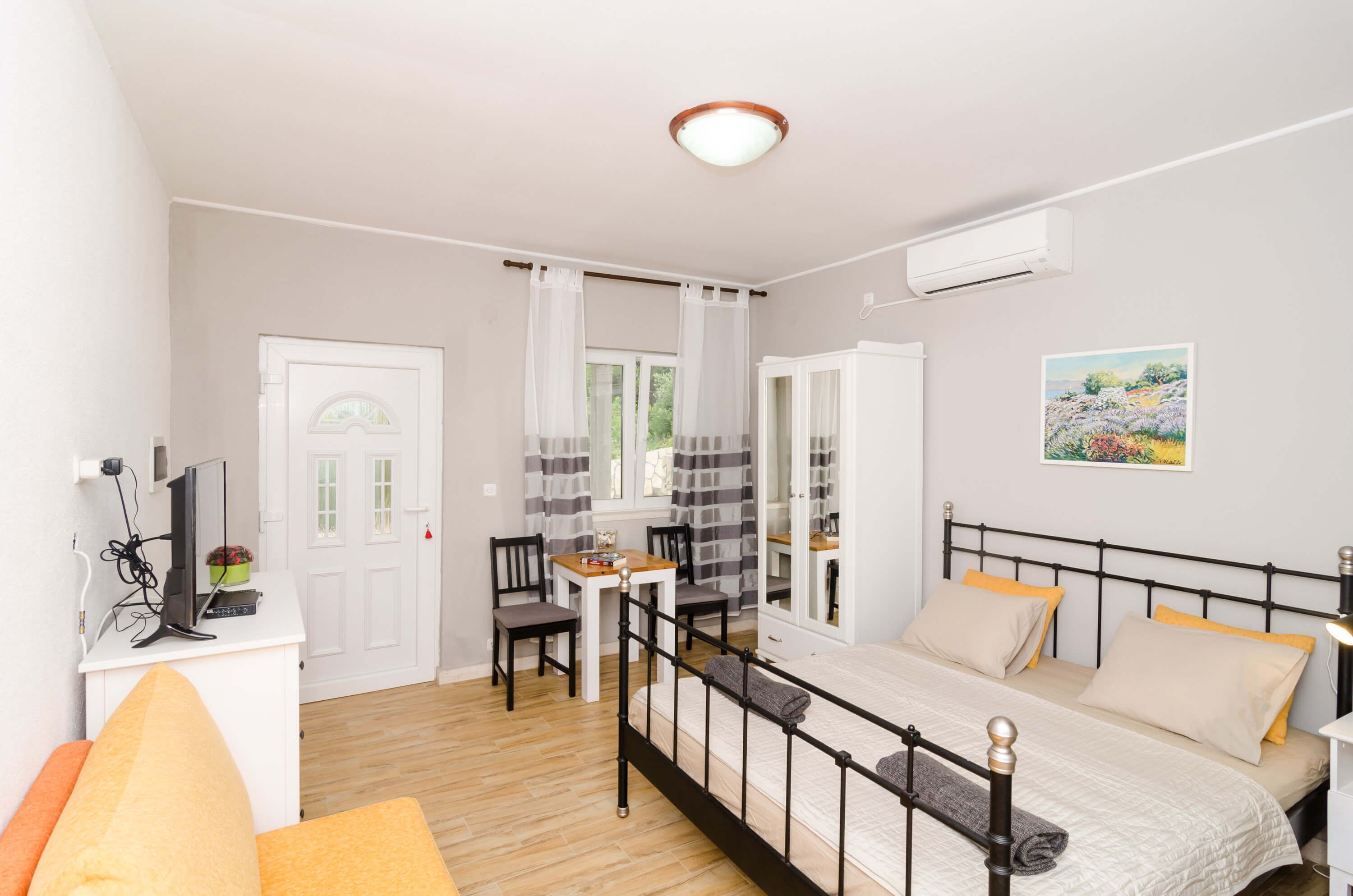 Ein Schlafzimmer mit einem Bett, Nachttisch und Schreibtisch mit Stuhl. Es gibt ein Fenster mit Vorhängen, eine Tür und ein Bild an der Wand.