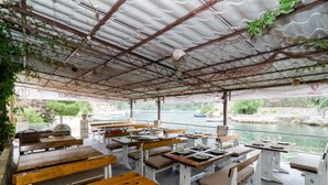 Un restaurante al aire libre con mesas y bancos de madera, techo de lámina corrugada y vistas a un río.