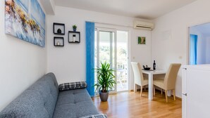 A modern living room with a grey sofa, a dining table with chairs, a potted plant, and a large window with blue curtains. - Mljet Gem in Pomena!! (Pomena)
