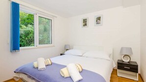 Una cama bien hecha con una colcha azul y dos toallas, una ventana con cortinas azules, y dos cuadros en la pared.