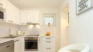 A modern kitchen with white cabinetry, a built-in oven, and a window with blinds. - White Stone Steel - 2 Bedroom Apartment with Terrace and Sea View (Dubrovnik)
