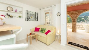 A cozy living room with a beige sofa, a small table, and a coffee table. There is a bookshelf with decorative items and a painting on the wall. A door leads to an outdoor area with a stone archway and a view of greenery. - White Stone Steel - 2 Bedroom Apartment with Terrace and Sea View (Dubrovnik)