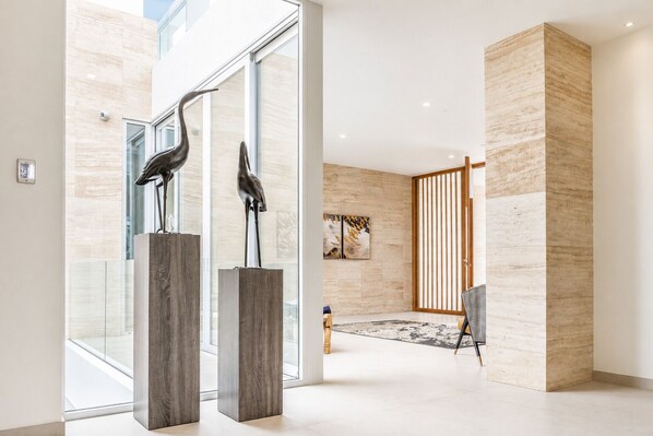 A modern interior with a glass door entrance, two sculptures on pedestals, a wooden partition, and a framed abstract painting.