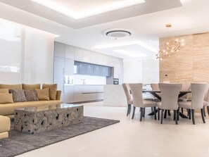 A modern living room with a sofa, coffee table, dining table with chairs, and a kitchen in the background.