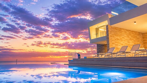 Ein modernes Gebäude mit Pool und Liegestühlen, mit Blick auf ein Gewässer bei Sonnenuntergang.