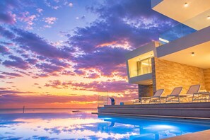 Un bâtiment moderne doté d’une piscine et de chaises longues, offrant une vue sur une étendue d’eau au coucher du soleil.