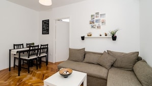 Un salon avec un canapé, une table à manger et des chaises, une table basse blanche et un luminaire suspendu. On y trouve également une étagère fixée au mur, ornée de plantes, et un tableau noir avec du texte.