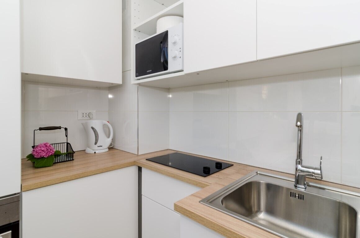 Une cuisine moderne avec des meubles blancs, un évier en acier inoxydable, une plaque de cuisson à induction noire, une bouilloire et un four à micro-ondes.