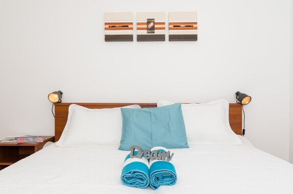 Ein ordentlich bezogenes Bett mit weißer und blauer Bettwäsche, ein Paar zusammengerollte Handtücher und ein an der Wand hängendes Kunstwerk.