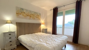 A bedroom with a wooden headboard, a large bed with white bedding, a nightstand with a lamp, and a view of the outdoors through a window with curtains. - 3* Apartment with terrace and parking, steps from Trestraou Beach in Perros-Guirec (Perros-Guirec)