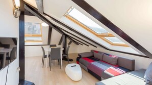 Ein Loft-Zimmer mit schrägen Wänden, Oberlichtern, einer Couch, einem Couchtisch und einem Essbereich mit Stühlen.