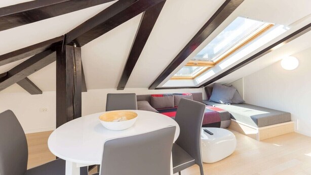 Ein modernes Loft-Schlafzimmer mit Dachfenster, einer Essecke mit weißem Tisch und Stühlen sowie einer gemütlichen Sitzecke mit Kissen.
