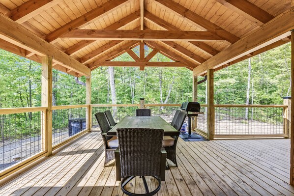 A covered outdoor deck with a wooden table and chairs, surrounded by a wooden railing, overlooking a forested area.