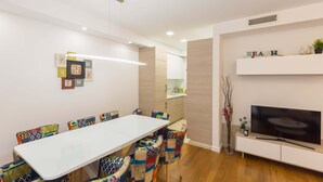 Un'area pranzo moderna con tavolo bianco e sedie colorate, un televisore a schermo piatto su un mobile bianco e una mensola con oggetti decorativi.