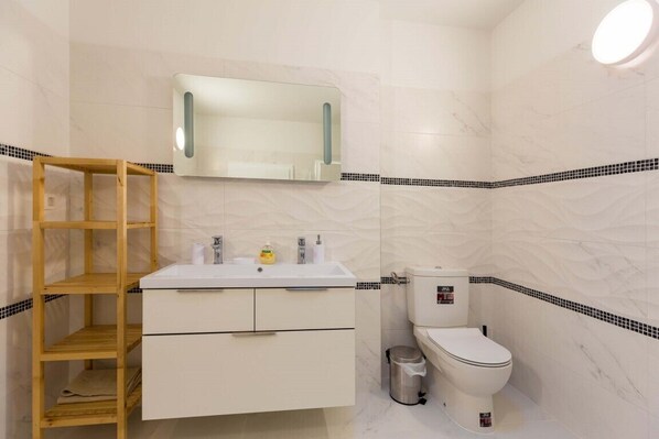A bathroom with a wooden shelf, a white sink, a mirror, a toilet, and a trash bin. - SOFIA One bedroom loft apartment 4pax (Dubrovnik)