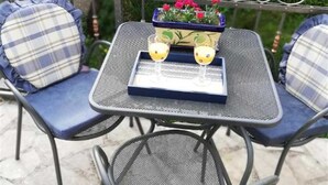 Une terrasse avec une table et des chaises en métal, deux verres contenant une boisson jaune et un petit pot de fleurs avec des fleurs rouges.