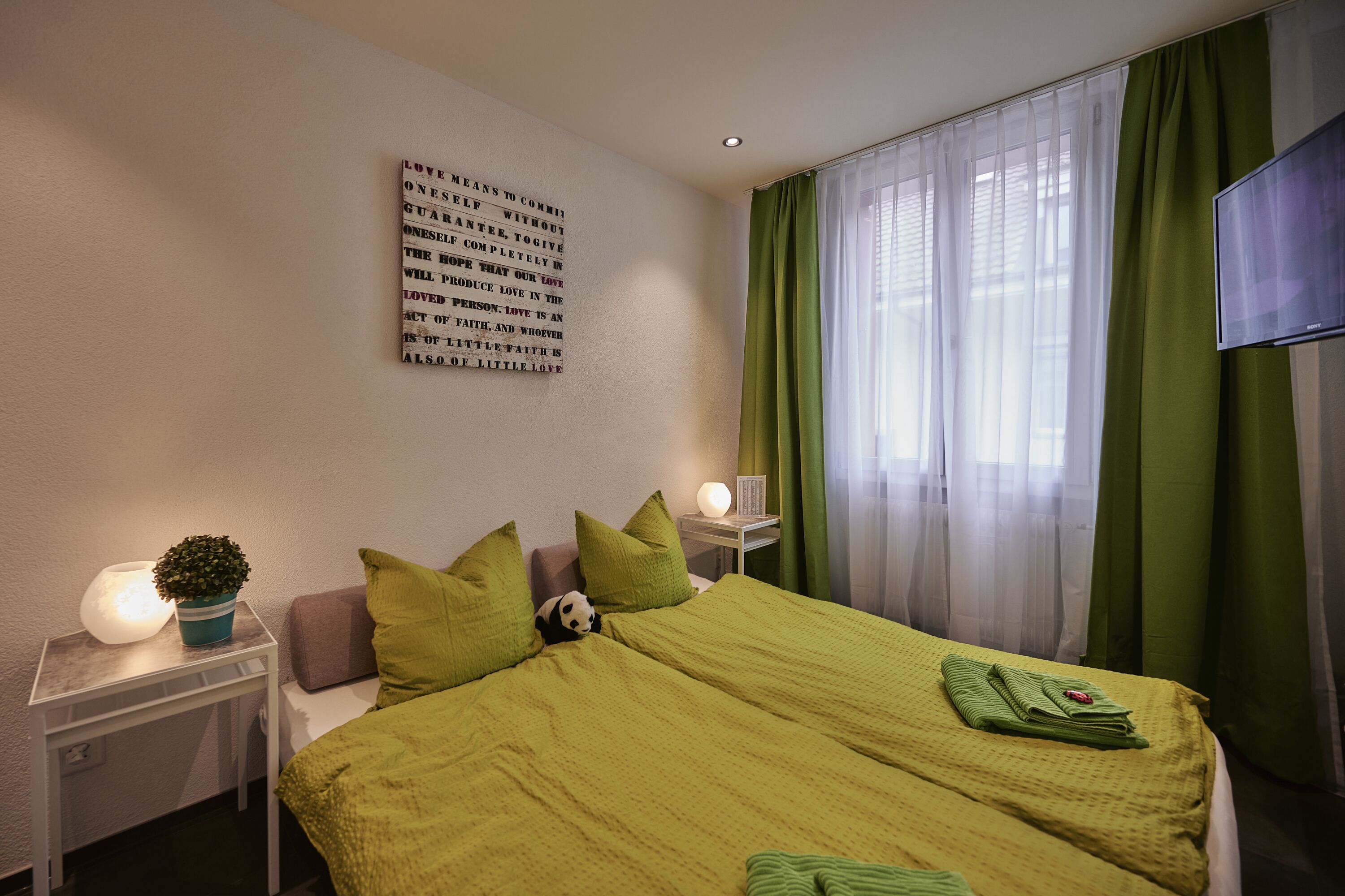 Ein ordentlich eingerichtetes Hotelzimmer mit Bett, grünen Vorhängen, einem an der Wand montierten Fernseher, einer Nachttischlampe und einem motivierenden Zitat an der Wand.