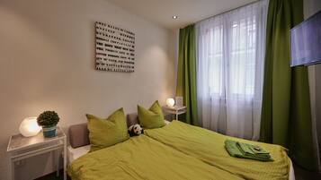 A neatly arranged hotel room with a bed, green curtains, a wall-mounted TV, a bedside lamp, and a motivational quote on the wall.
