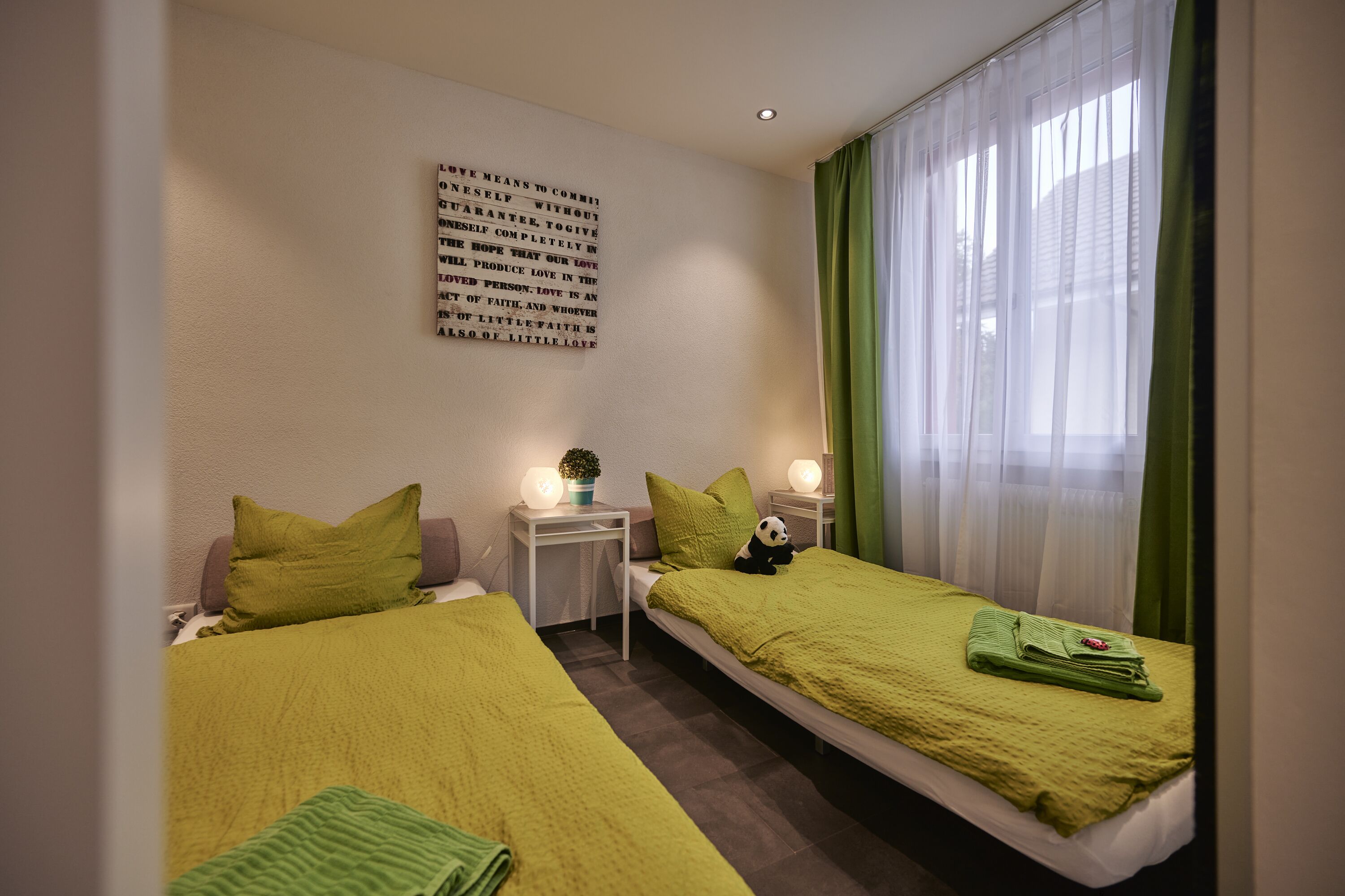 A hotel room with two beds, a wall-mounted light fixture, and a window with sheer curtains.