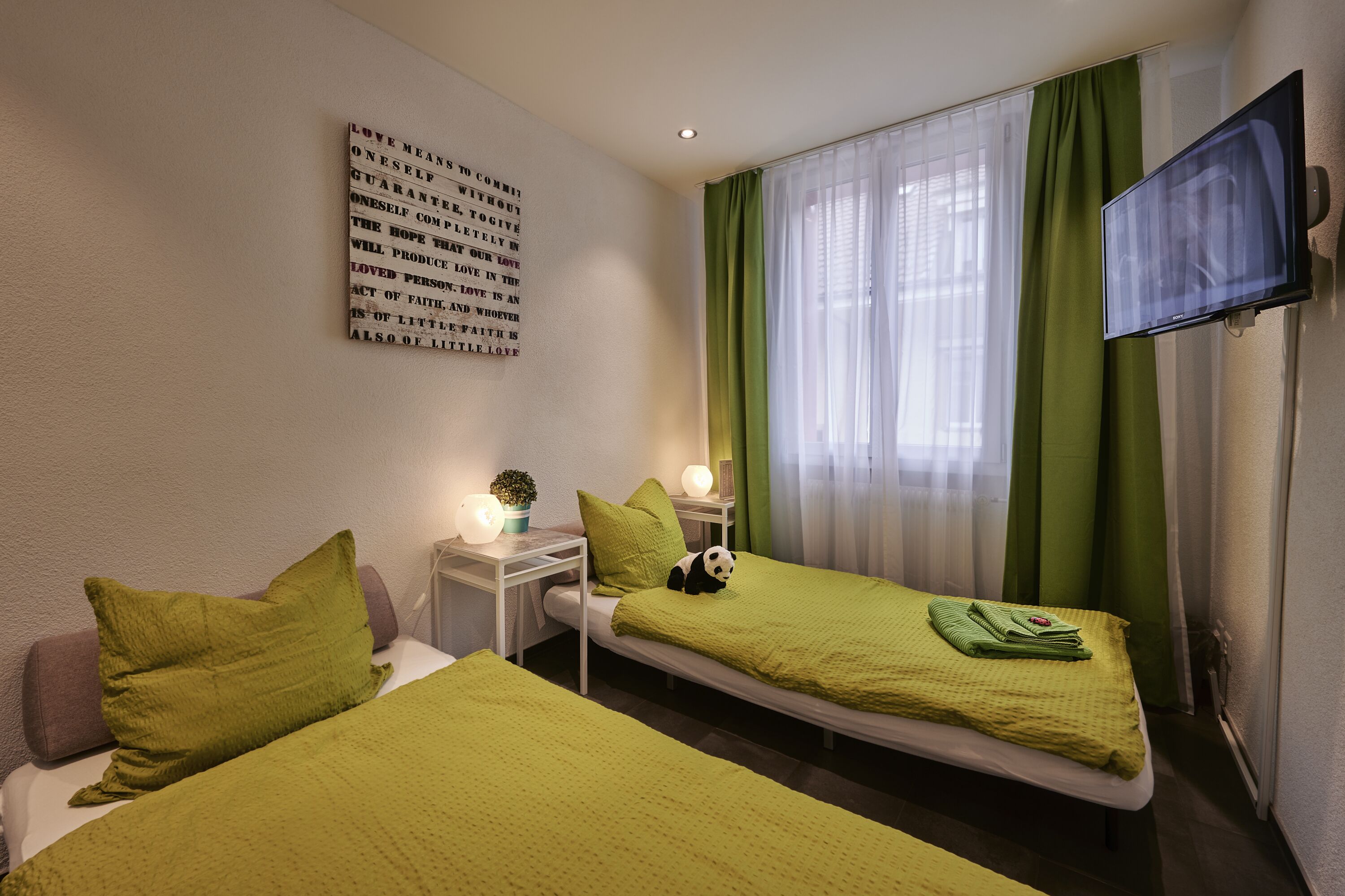 A hotel room with two single beds, a flat-screen TV, a wall-mounted TV, a bedside table with a lamp, and a wall with a motivational quote.