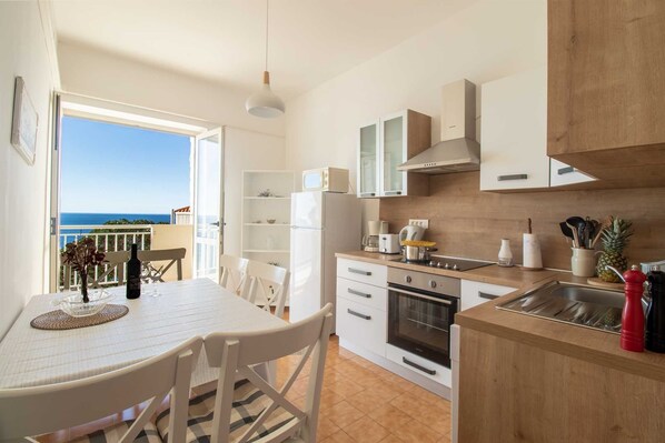 A modern kitchen with a view of the sea, featuring white cabinetry, a wooden countertop, and basic appliances.
