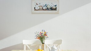 Una mesa de comedor con mantel blanco, un florero de flores, un bol de fruta, una tetera blanca y una jarra blanca.