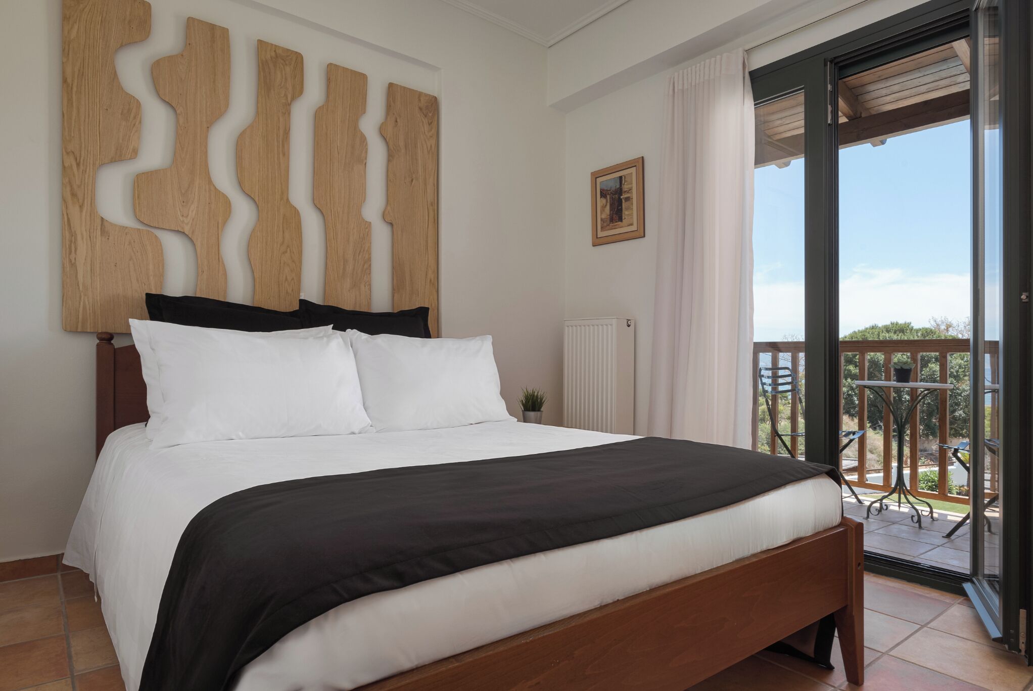 Camera da letto con un grande letto, una testata in legno, vista sull'esterno tramite una porta scorrevole in vetro e un piccolo tavolo con sedie all'esterno.