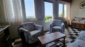 Una habitación con dos sillones, un sofá, una mesa de centro de madera y un suelo a cuadros. Hay dos ventanas con cortinas, un armario de madera con una cama, y un cuadro en la pared.