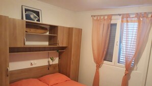 Un dormitorio con cama, armario de madera, repisa con un cuadro y ventana con cortinas.
