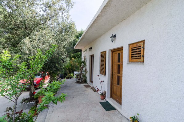Un étroit sentier mène à un bâtiment blanc doté de portes et de volets en bois. Des plantes en pot et une voiture rouge sont disposées le long du sentier.