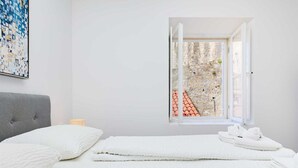 Ein Schlafzimmer mit Bett, Kissen, einer Nachttischlampe und einem Fenster mit Blick auf eine Steinsäule und rote Kacheln.
