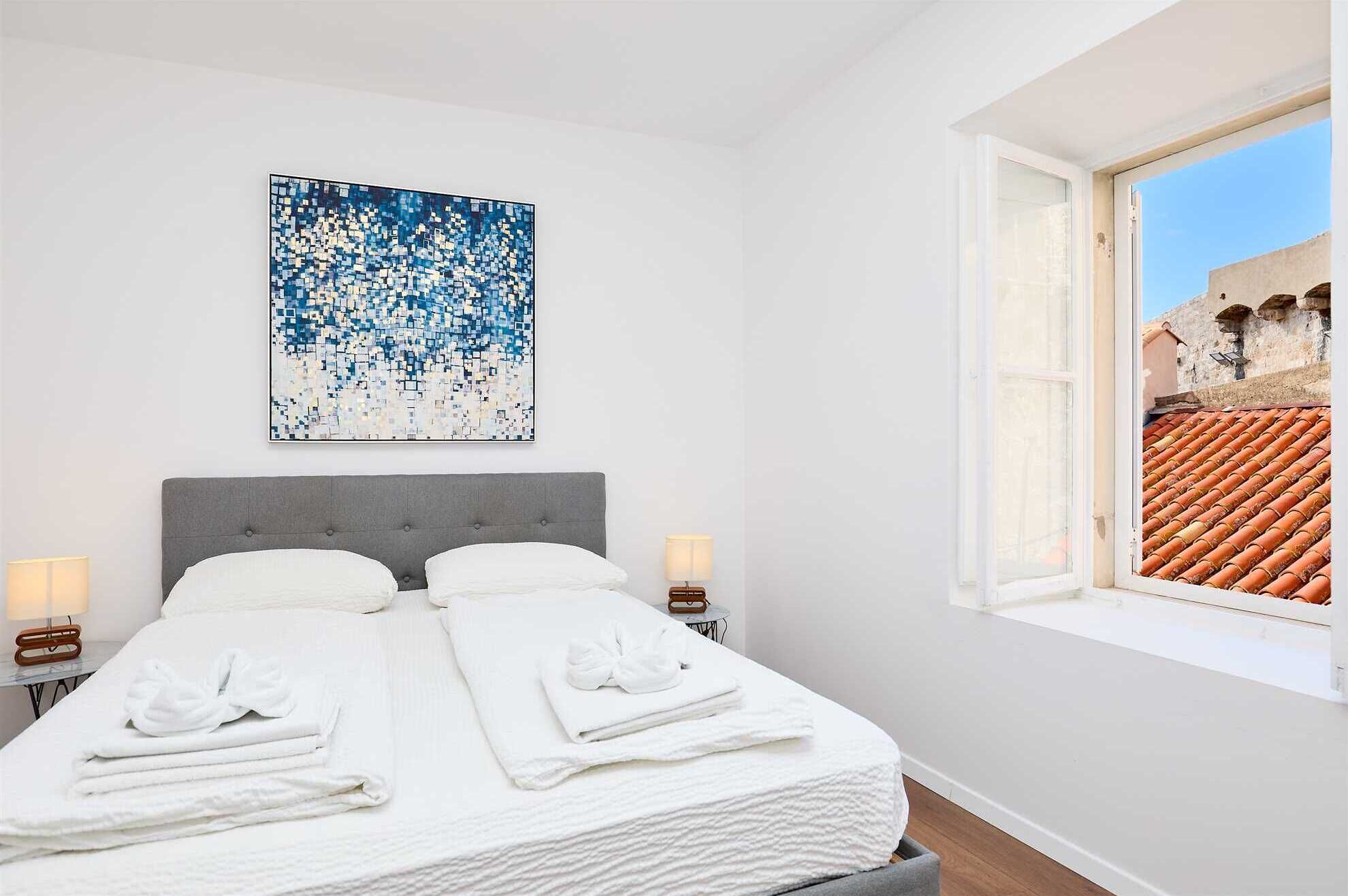 Een hotelkamer met een bed, twee nachtkastjes, een raam met uitzicht op een dak met rode tegels, en een abstract schilderij in blauw en wit aan de muur.