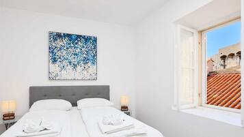Een hotelkamer met een bed, twee nachtkastjes, een raam met uitzicht op een dak met rode tegels, en een abstract schilderij in blauw en wit aan de muur.