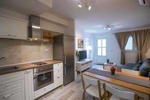 Une cuisine moderne avec des meubles blancs, un four encastré et un réfrigérateur en acier inoxydable. On y trouve un coin repas avec une table et des chaises en bois, ainsi qu’un salon avec un canapé et un téléviseur.