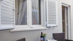 Un balcón con sillas de mimbre, una mesa con una botella de vino y una ventana con contraventanas blancas.