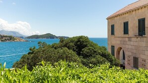 Un bâtiment situé sur la côte, doté d’une façade en pierre et de volets verts, qui offre une vue imprenable sur une mer bleue et limpide et une végétation luxuriante.