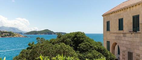 A coastal building with a stone facade and green shutters, overlooking a clear blue sea and lush greenery.