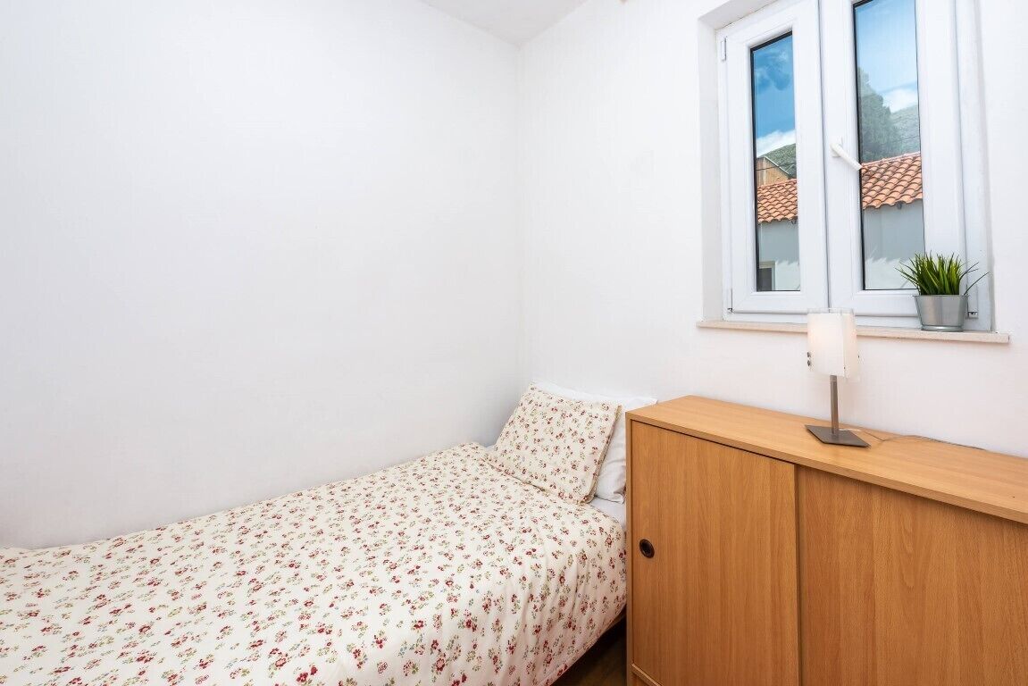 A small, neatly arranged bedroom with a single bed, a wooden nightstand, a bedside lamp, and a window with a view of a tiled roof.