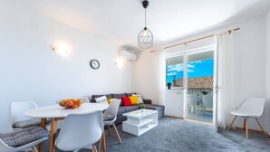 Ein modernes Wohnzimmer mit Sofa, Esstisch und Stühlen. Ein Balkon bietet Blick auf Himmel und Häuser.
