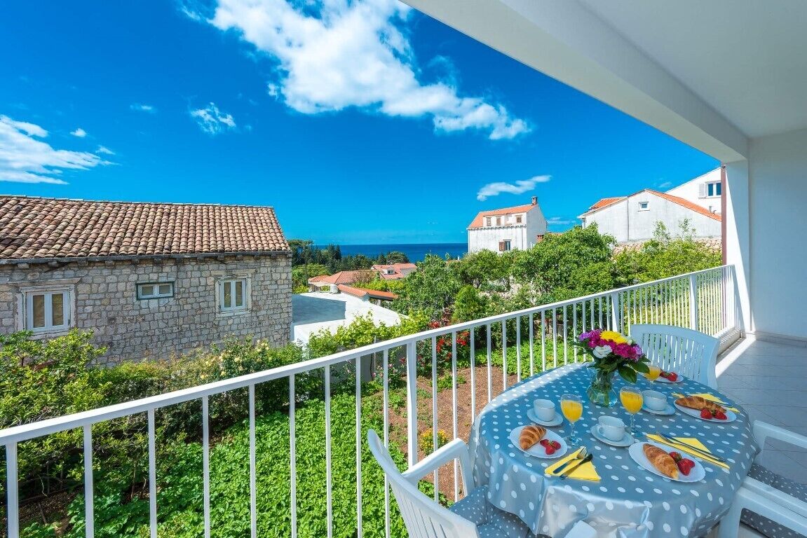 A balcony with a table set for breakfast, overlooking a coastal town with stone buildings and a clear blue sky.