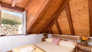 A bedroom with a wooden ceiling, a bed with a white and pink pillow, a bedside table with a vase of flowers, and a window overlooking a village. - Daniela Old Town three bedroom apartment (Dubrovnik)