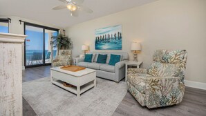A living room with a white coffee table, a grey sofa with teal pillows, a floral armchair, a painting of a coastal scene, and a ceiling fan.
