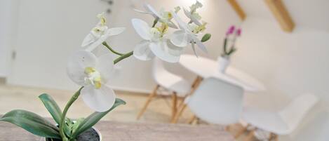 Una pianta di orchidea in vaso su un tavolo di legno, in una stanza con una sedia bianca e un tavolino rotondo sullo sfondo.
