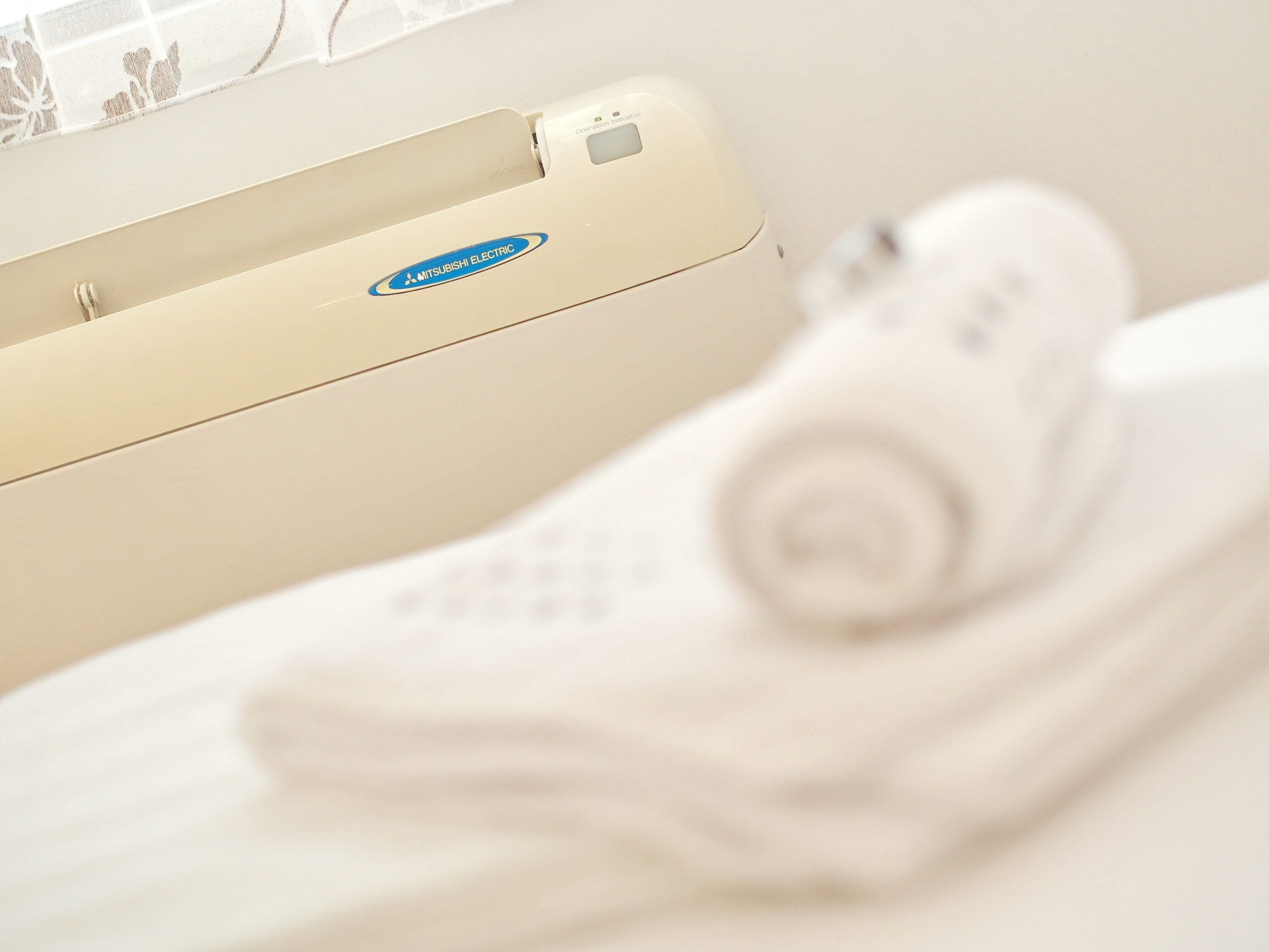 Un oreiller blanc et une couverture blanche sur un lit, avec un mur de couleur beige et une fenêtre ornée d’un rideau blanc.