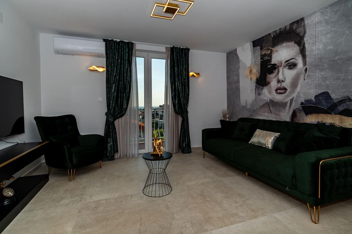Un salon moderne avec un canapé vert, un fauteuil noir et une petite table d’appoint. On y trouve une grande fenêtre avec des rideaux, un téléviseur à écran plat et un climatiseur fixé au mur.