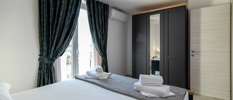 Quarto de hotel com cama, cortinas escuras, guarda-roupa e vista pela janela.