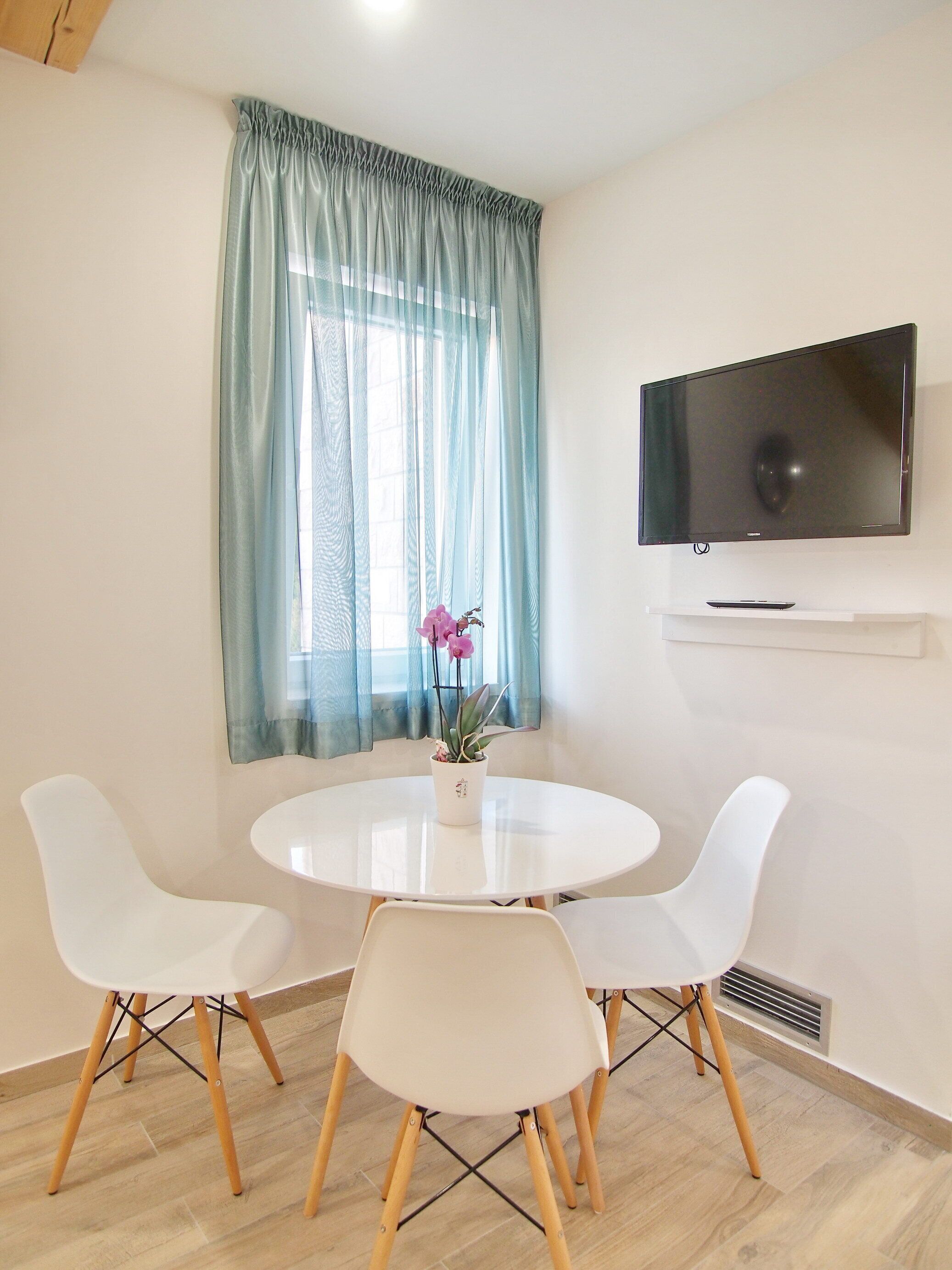 A modern room with a round white table, two white chairs, a flat-screen TV mounted on the wall, and a window with sheer curtains.