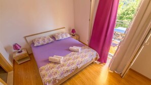 Un dormitorio con piso de madera, cama con colcha morada y almohadas florales, mesita de noche con lámpara, y vista al exterior a través de una ventana con cortinas rosadas.