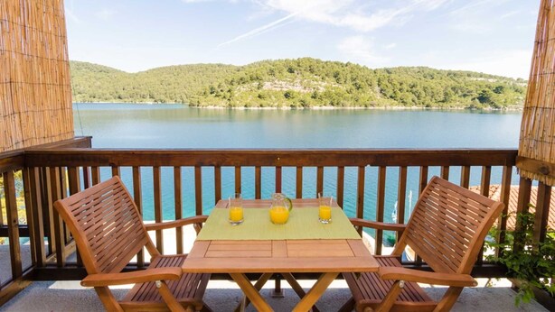 Une terrasse en bois avec une table et des chaises, donnant sur un lac, avec une colline boisée en arrière-plan.