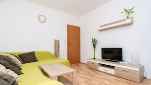 Un salon moderne avec un canapé vert, une table basse en bois, un téléviseur posé sur un meuble en bois et une petite plante sur une étagère.